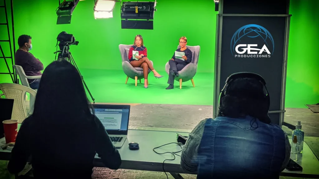 Técnico de GEA Producciones monitorizando grabación en estudio croma desde un portátil, con presentadoras al fondo y logo de GEA en pantalla.