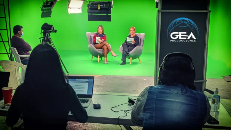 Técnico de GEA Producciones monitorizando grabación en estudio croma desde un portátil, con presentadoras al fondo y logo de GEA en pantalla.